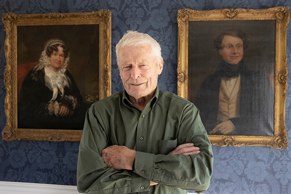 Richard Cocks standing between two oil paintings at his home.