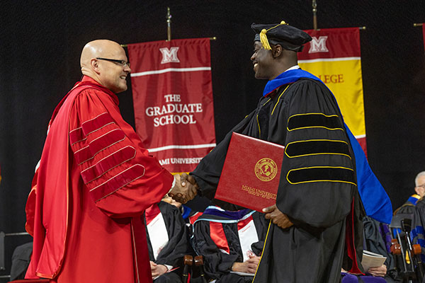 A doctoral candidate in academic regalia holds his degree and shakes the hand of President Greg Crawford at Fall Commencement ceremony