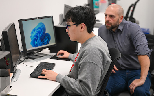 John Femiani, professor of Computer Science and Software Engineering, looks on as research assistant Ethan Han works at a computer. Han is one of two graduate students assisting Femiani on a project that uses artificial intelligence to enhance the detection of underwater mines.