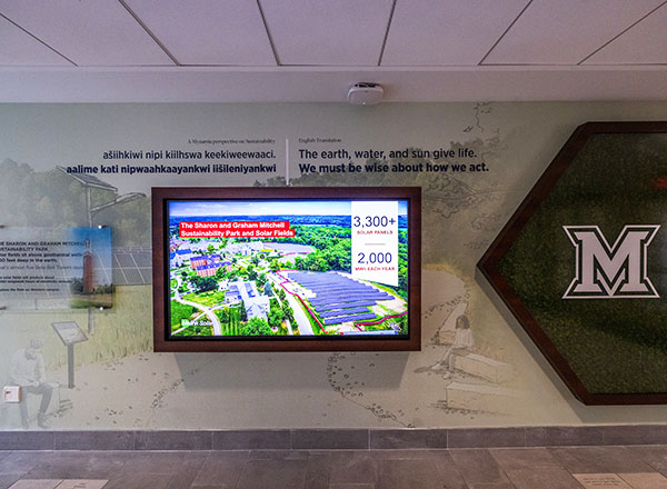 sustainability wall in armstrong student center with large graphic of park, a large digital monitor displaying a slide with miami's solar fields, and a large beveled M on a background of preserved moss